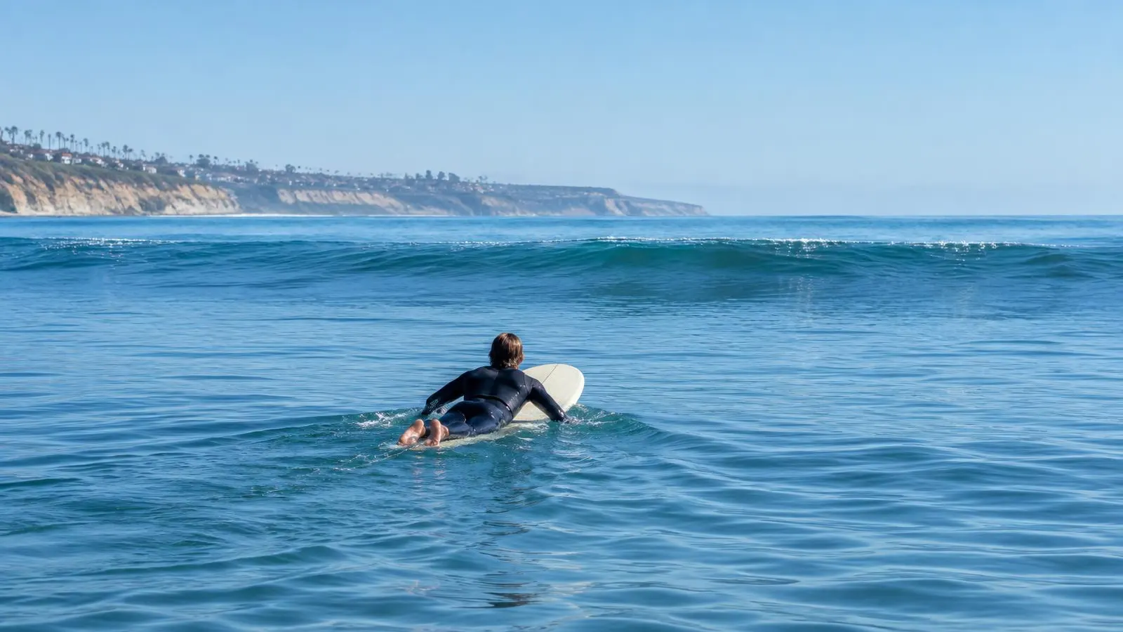 Surfer paddling a longboard through calm blue water toward small waves on a bright morning off the North County coast.